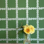 A vase of flowers stands in front of a wall covered in a floral grid print in white on a green field.