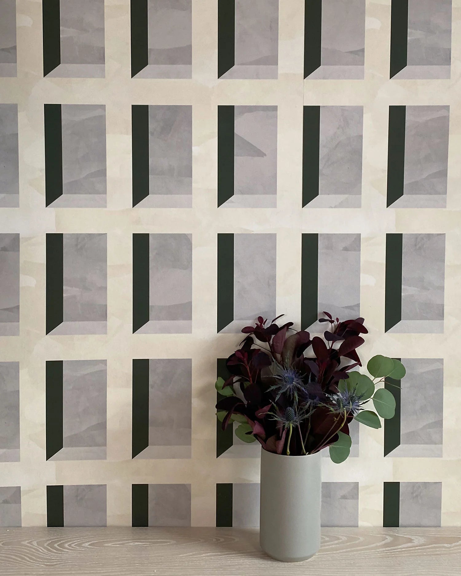A vase of flowers stands in front of a wall papered in a dimensional grid print in shades of cream, gray and charcoal.