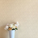 A vase of flowers stands in front of a wall papered in a painterly leaf print in tan and white on a cream field.