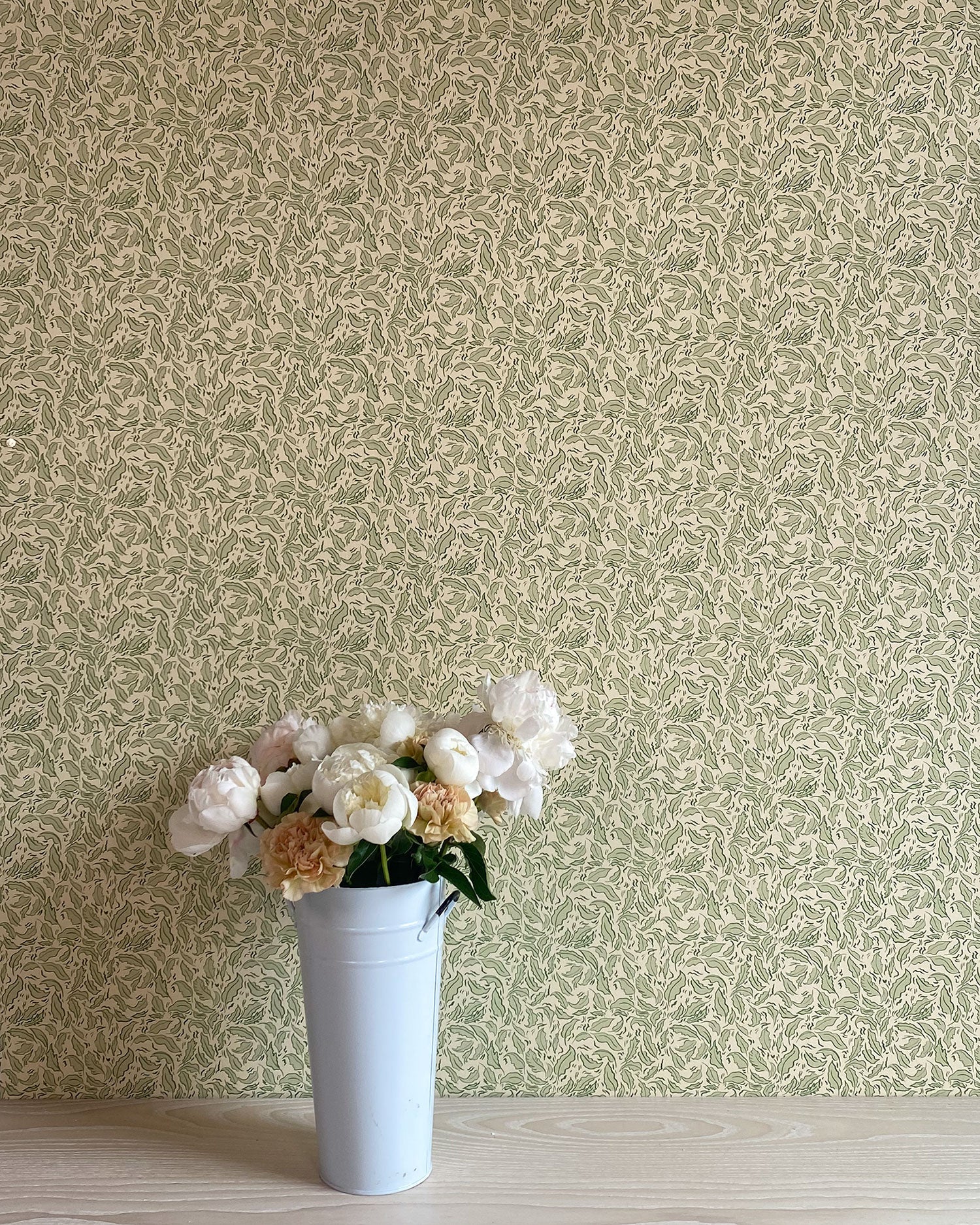 A vase of flowers stands in front of a wall papered in a painterly leaf print in olive and black on a cream field.