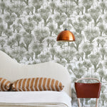 A modernist bed, hanging lamp and chair stand in front of a wall papered in a painterly tree print in sage and white.