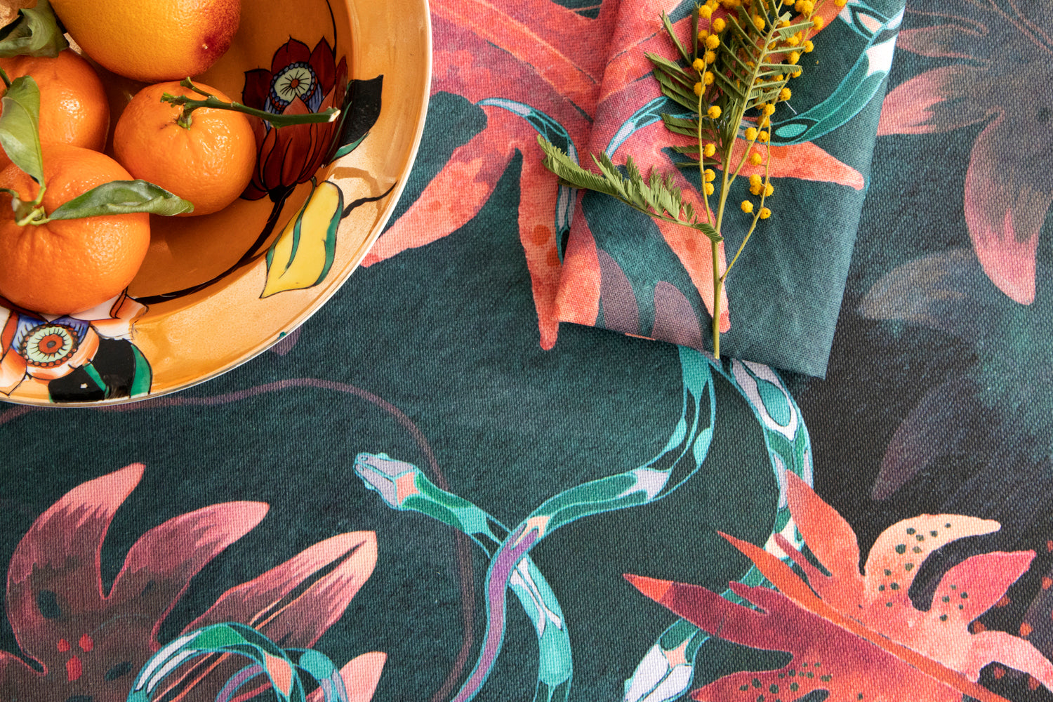 Detail of tablecloth vignette in a playful leaf and snake print in pink, blue and green on a dark turquoise field.