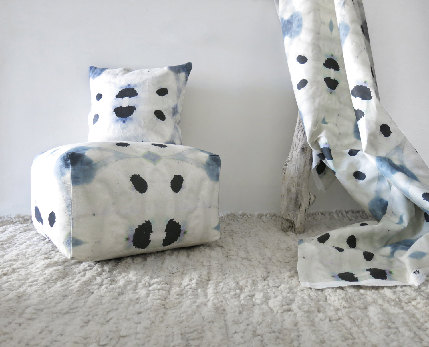 Tableau with draped fabric, pillows and sqaure pouffe in a watercolor ink blot print in shades of charcoal and black on a tan field.