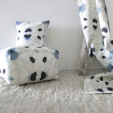 Tableau with draped fabric, pillows and sqaure pouffe in a watercolor ink blot print in shades of charcoal and black on a tan field.