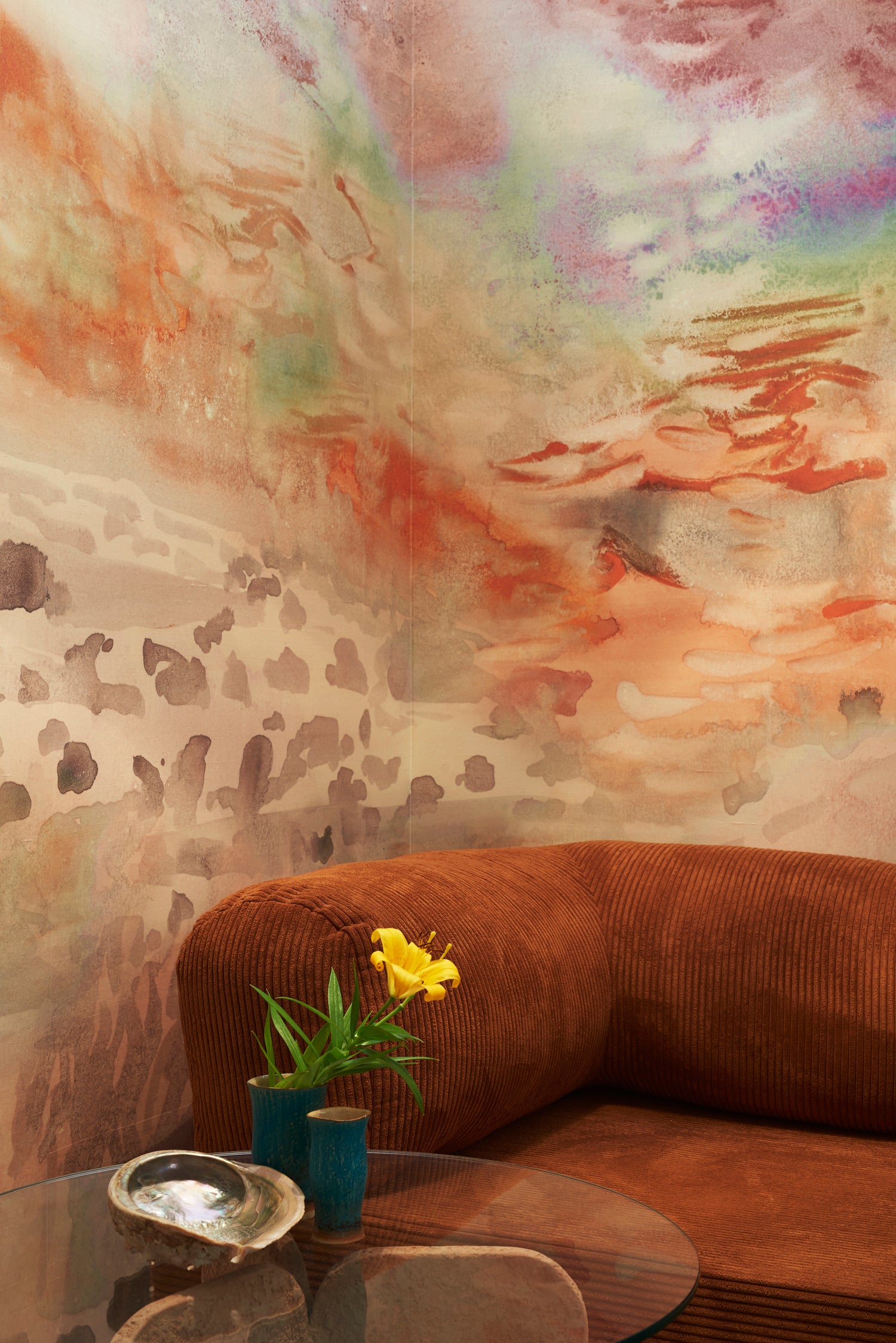 Living room with modernist couch and pendant lamp against ethereal watercolor mural in rust red, warm orange, tan, and taupe.