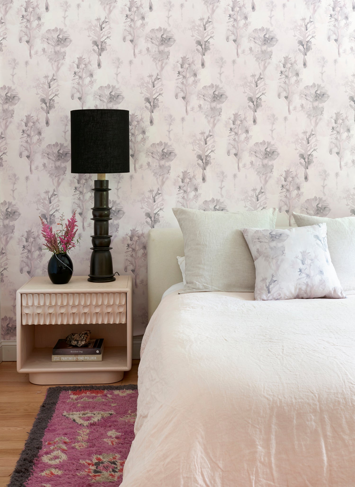 Bedroom with a white linened bed, side table and agaist a watercolor tree print in light grey and pink. 