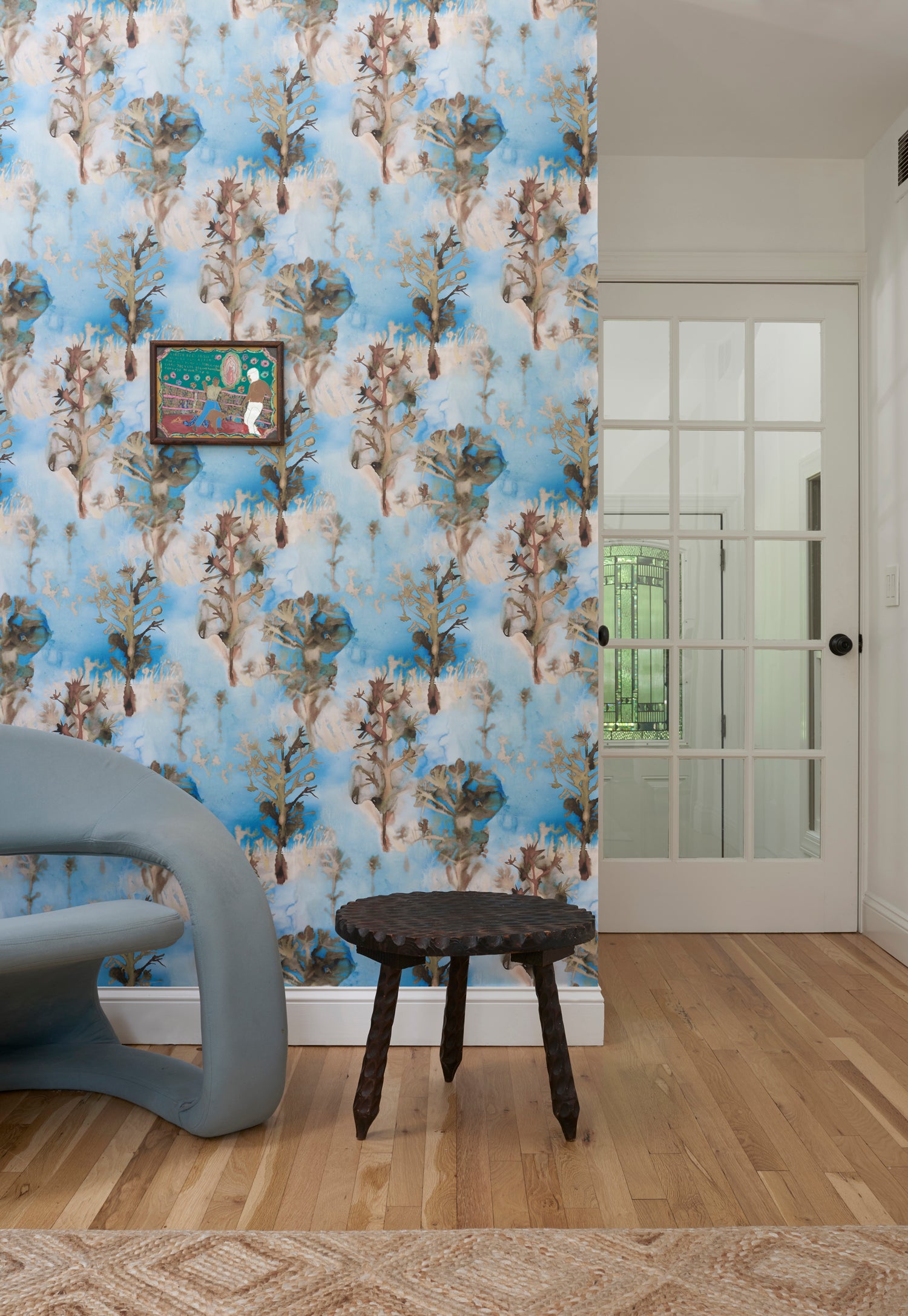 Living room interior with blue accent chair against wallpaper in watercolor tree print in shades of tan and gray on a blue field.