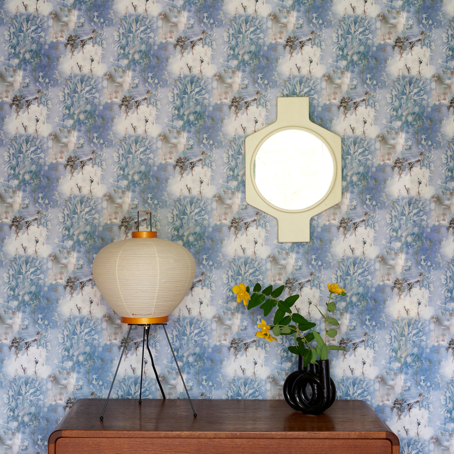 Wooden credenza with paper lantern and props against in a painterly tree print in blue and grey. 