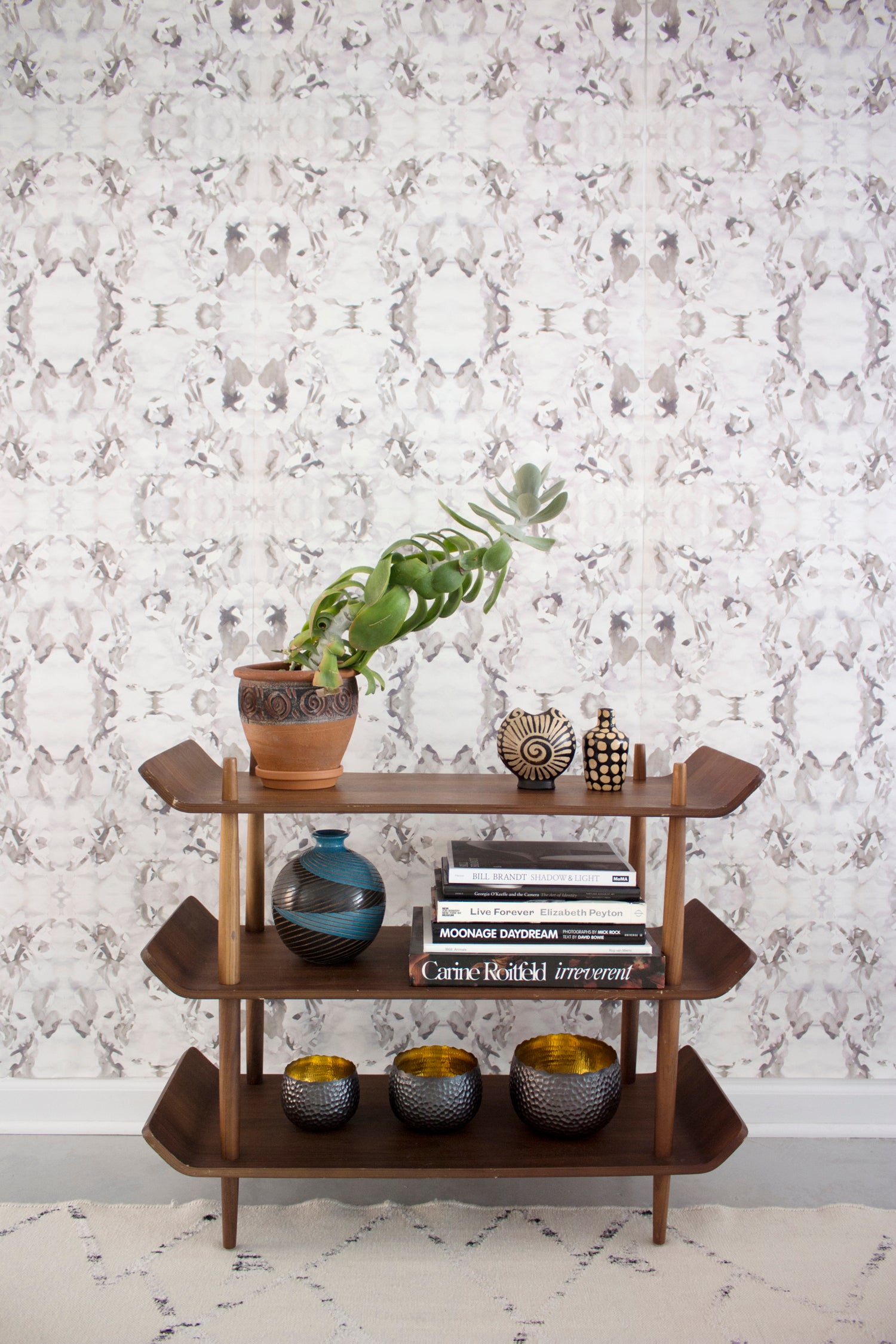 Wallpaper install in a watercolor paint blotch print in purple and gray on a white field with a wooden bookshelf with accessories.