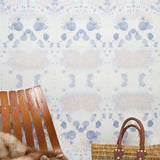 Wallpaper install detail in a watercolor inkblot print in pastel pink and purple on a cream field with a wooden slat chair and woven bag. 
