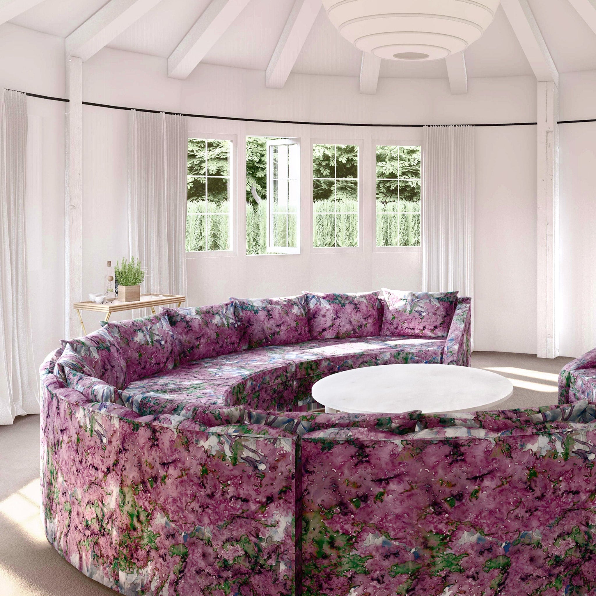 A large round sofa upholstered in a painterly tree print in purple, green and brown stands in the middle of a sunny room.