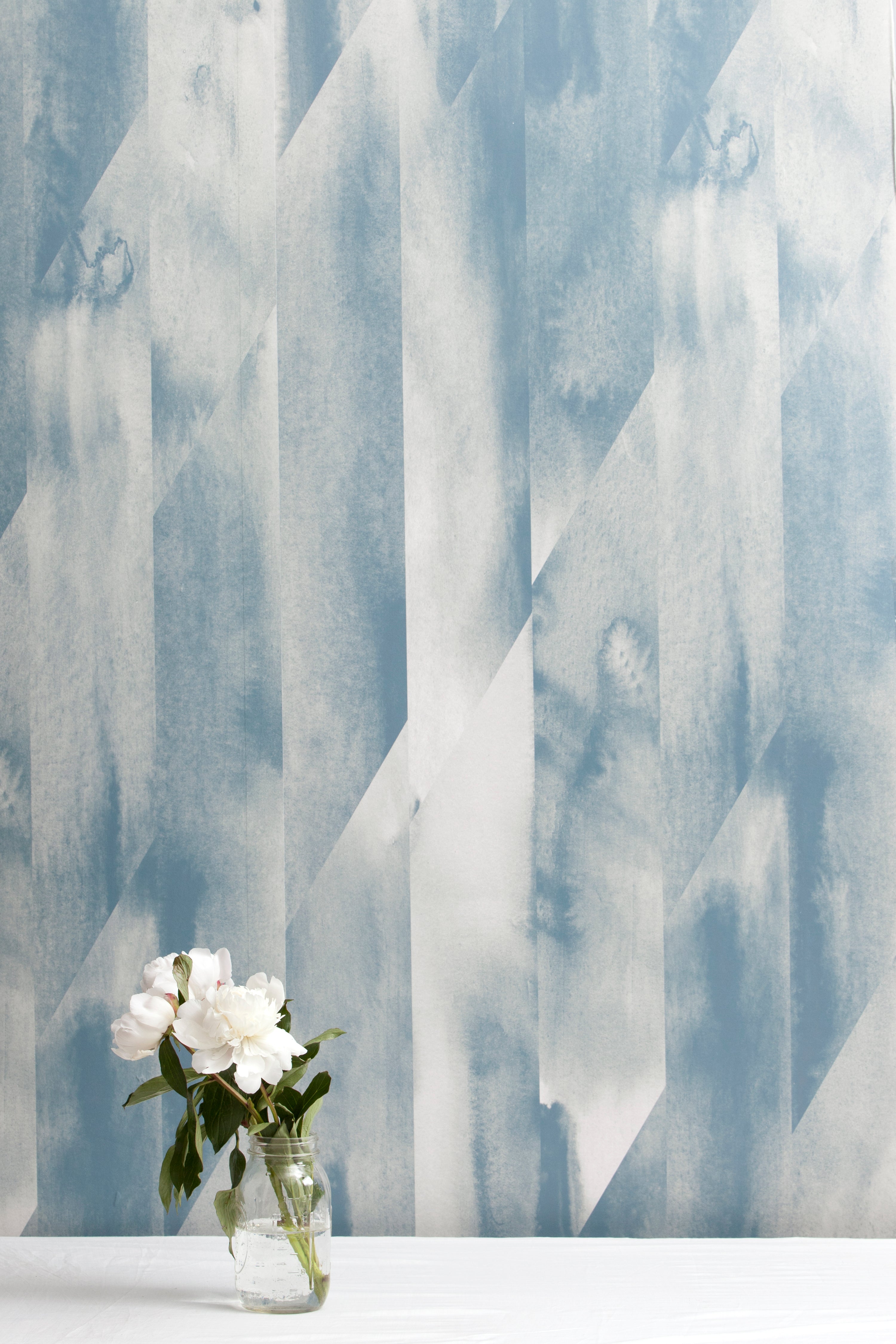 A vase of flowers stands in front of a wall papered in a linear geometric print in shades of blue and gray.