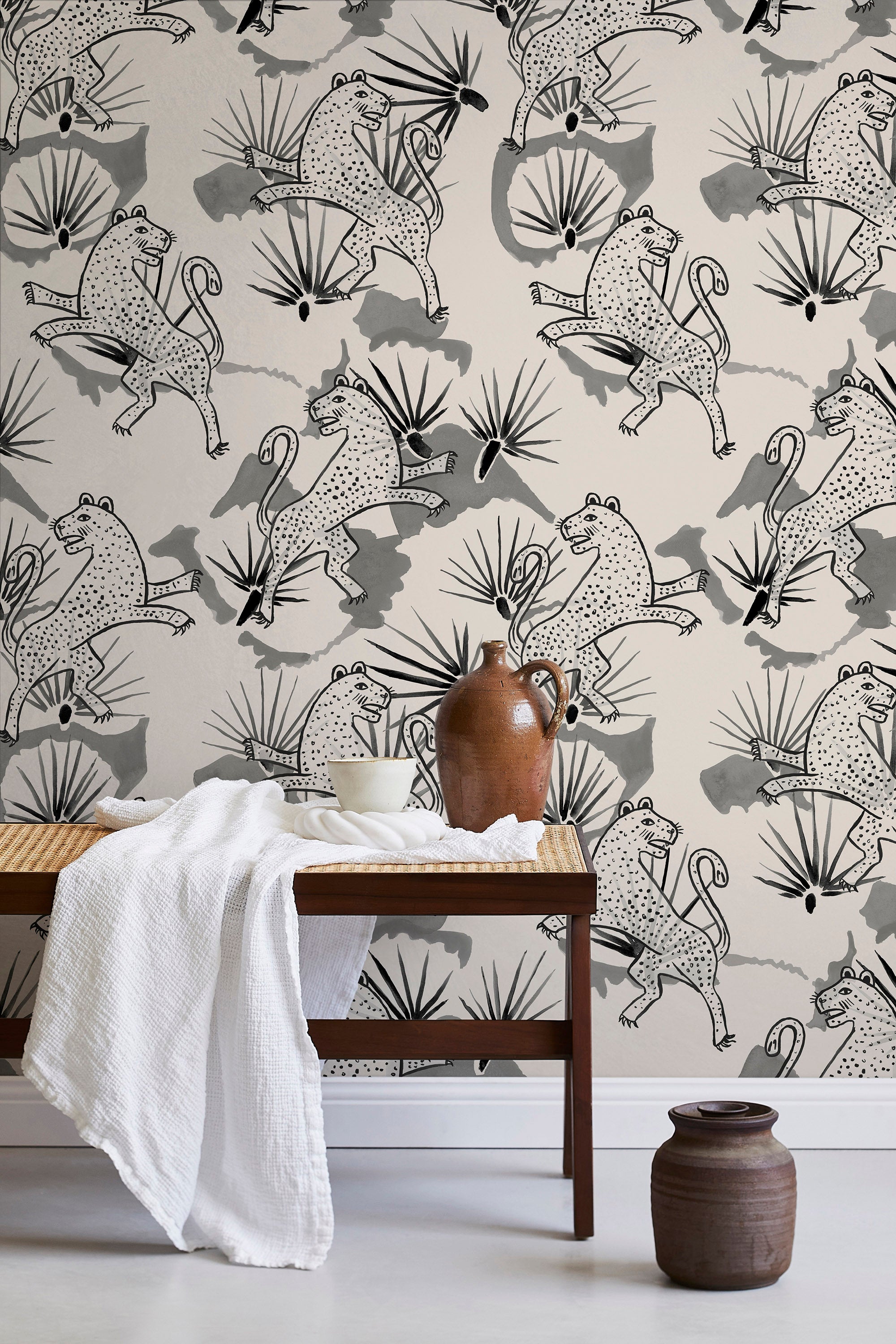 A bench with a towel and clay vases stands in front of a wall papered in a leopard and leaf print in gray and cream.