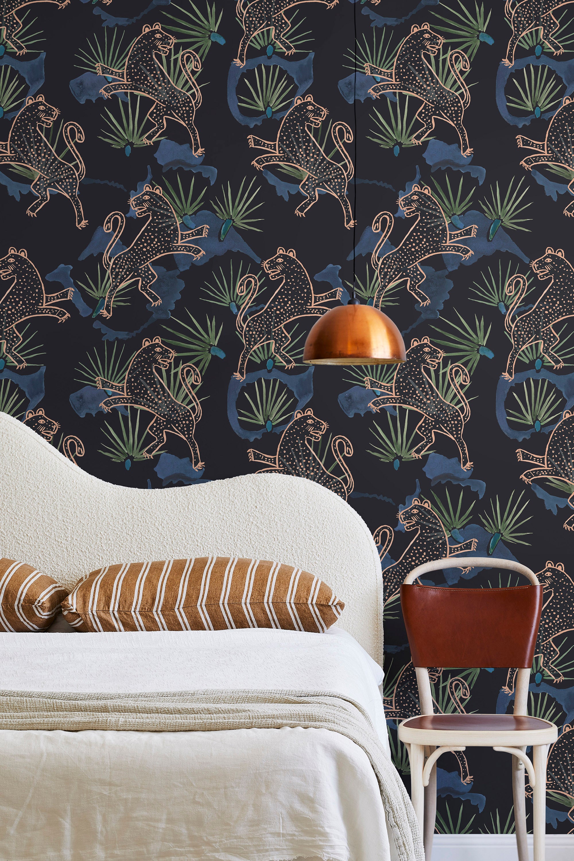 A modernist bed, hanging lamp and chair stand in front of a wall papered in a leopard and leaf print in pink and blue.