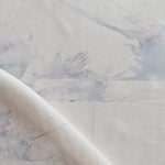 Draped fabric yardage in a watercolor splatter print in pastel purple and gray on a cream field.