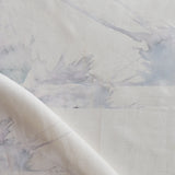 Draped fabric yardage in a watercolor splatter print in pastel purple and gray on a cream field.