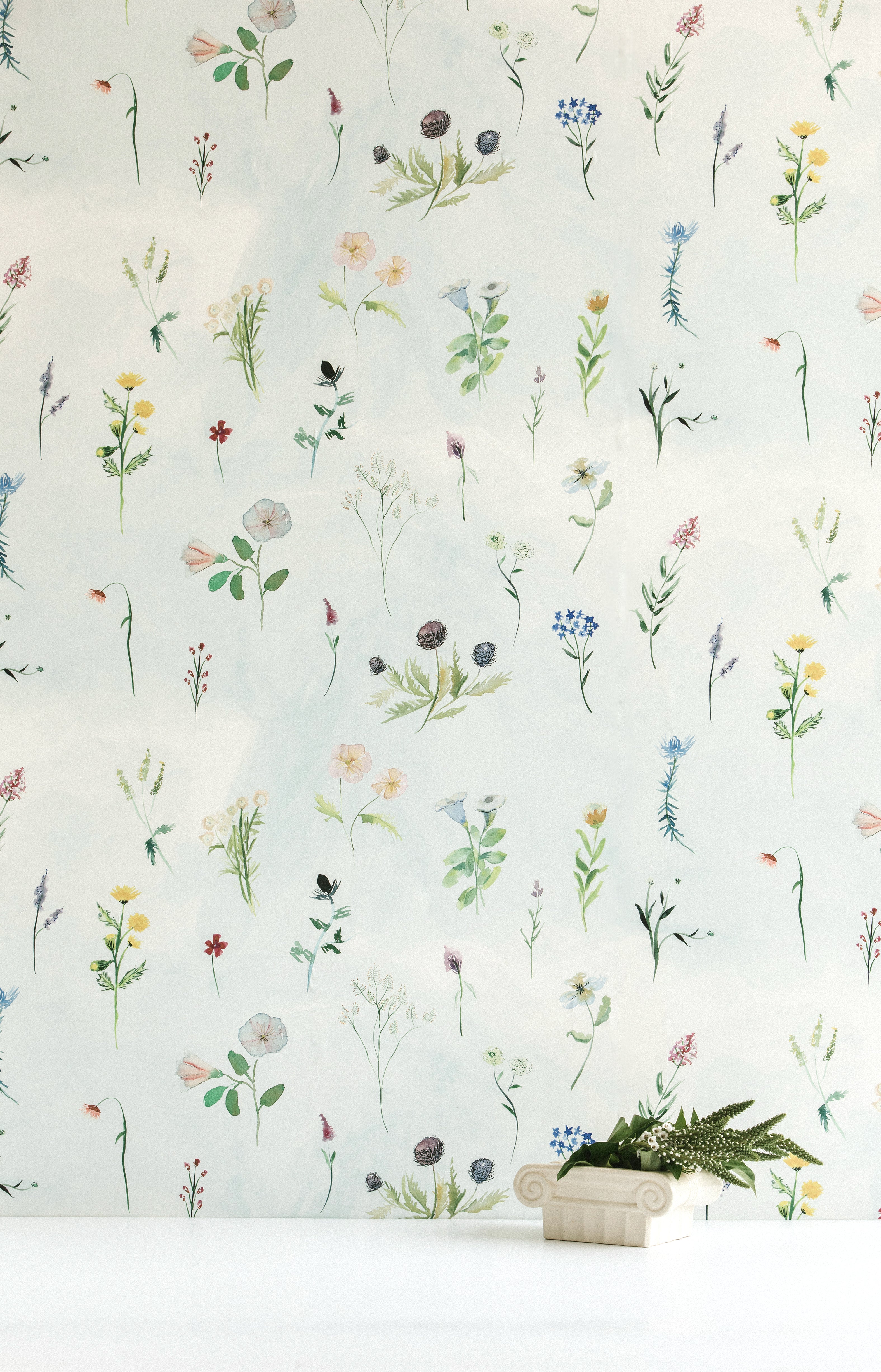 A vase of flowers stands in front of a wall covered in a painterly flower print in green and pastels on a cream field.