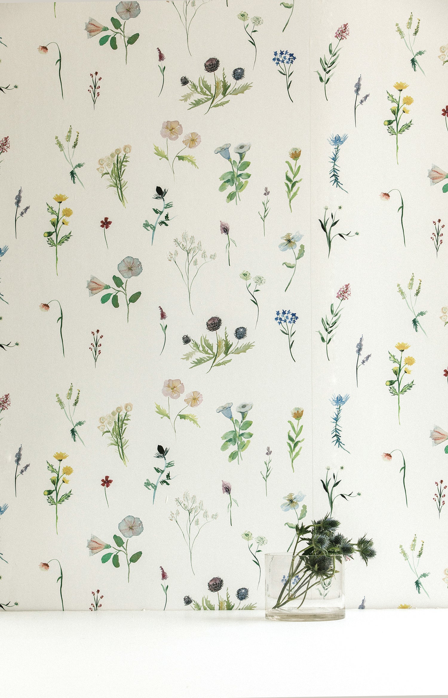 A vase of flowers stands in front of a wall covered in a painterly flower print in green and pastels on a cream field.