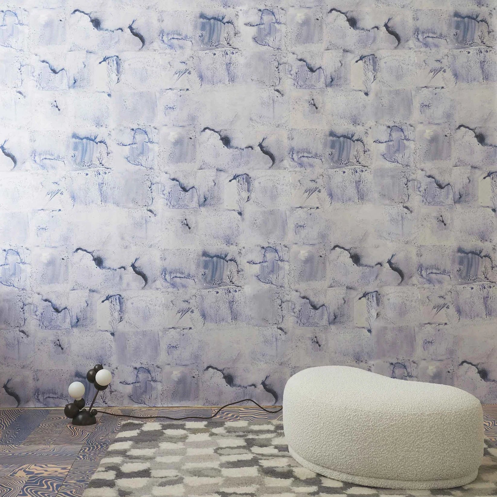 Room interior with a watercolor check wallpaper in blue featuring a white ottoman and small lamp.