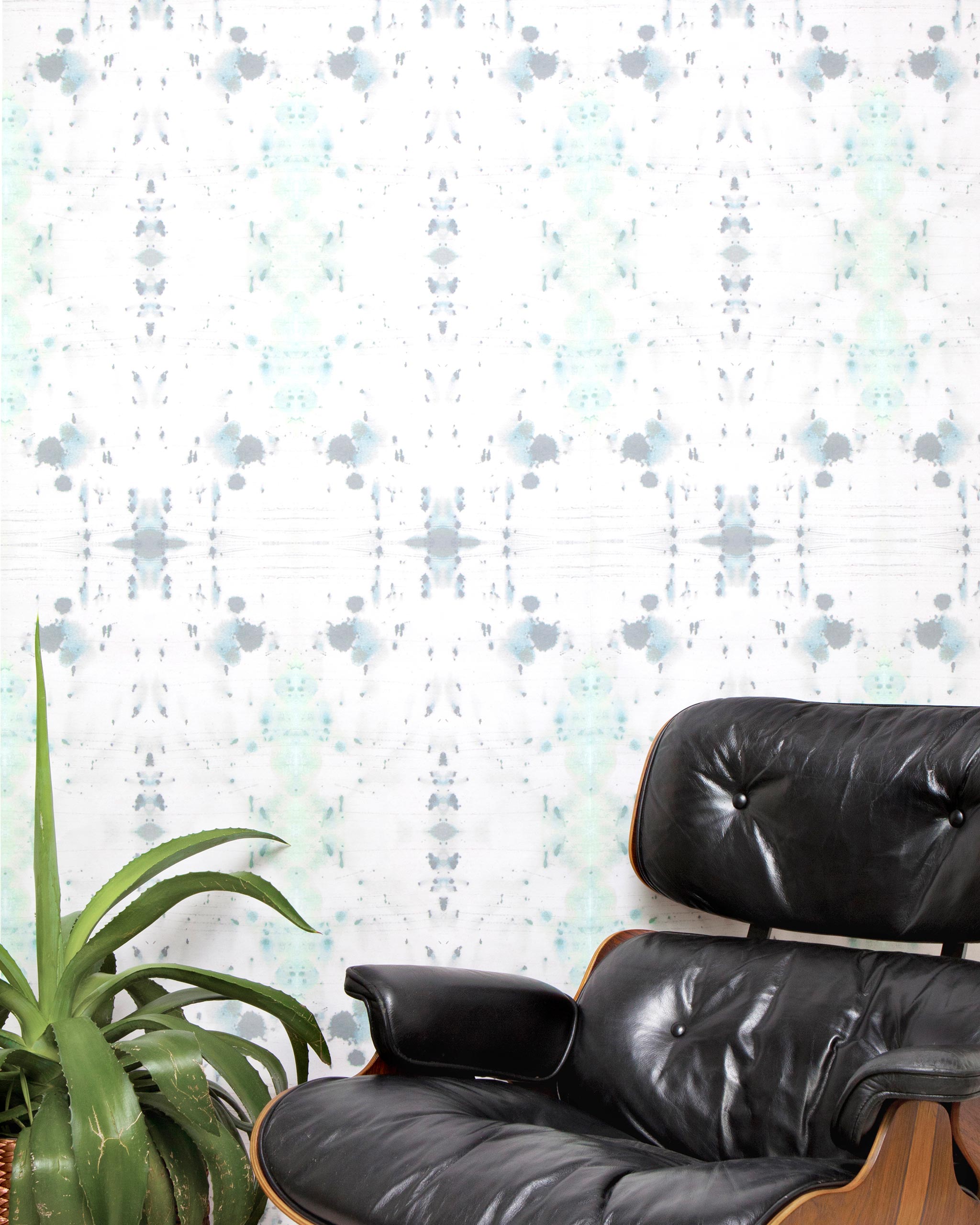 A chair and plant stand in front of a wall papered in a watercolor ink blot print in gray and green on a cream field.