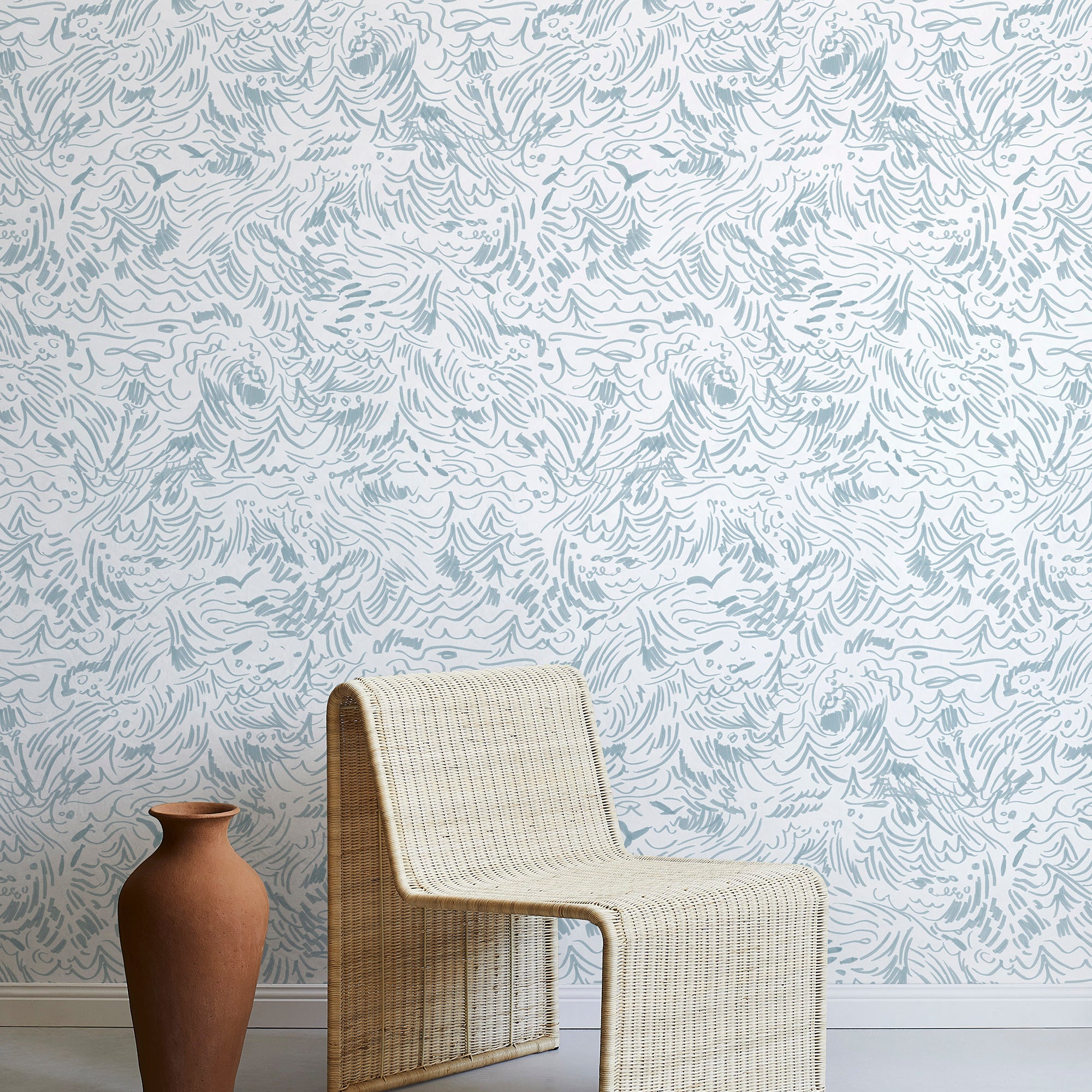 A straw chair and clay pot stand in front of a wall papered in a painterly wave print in light blue and white.