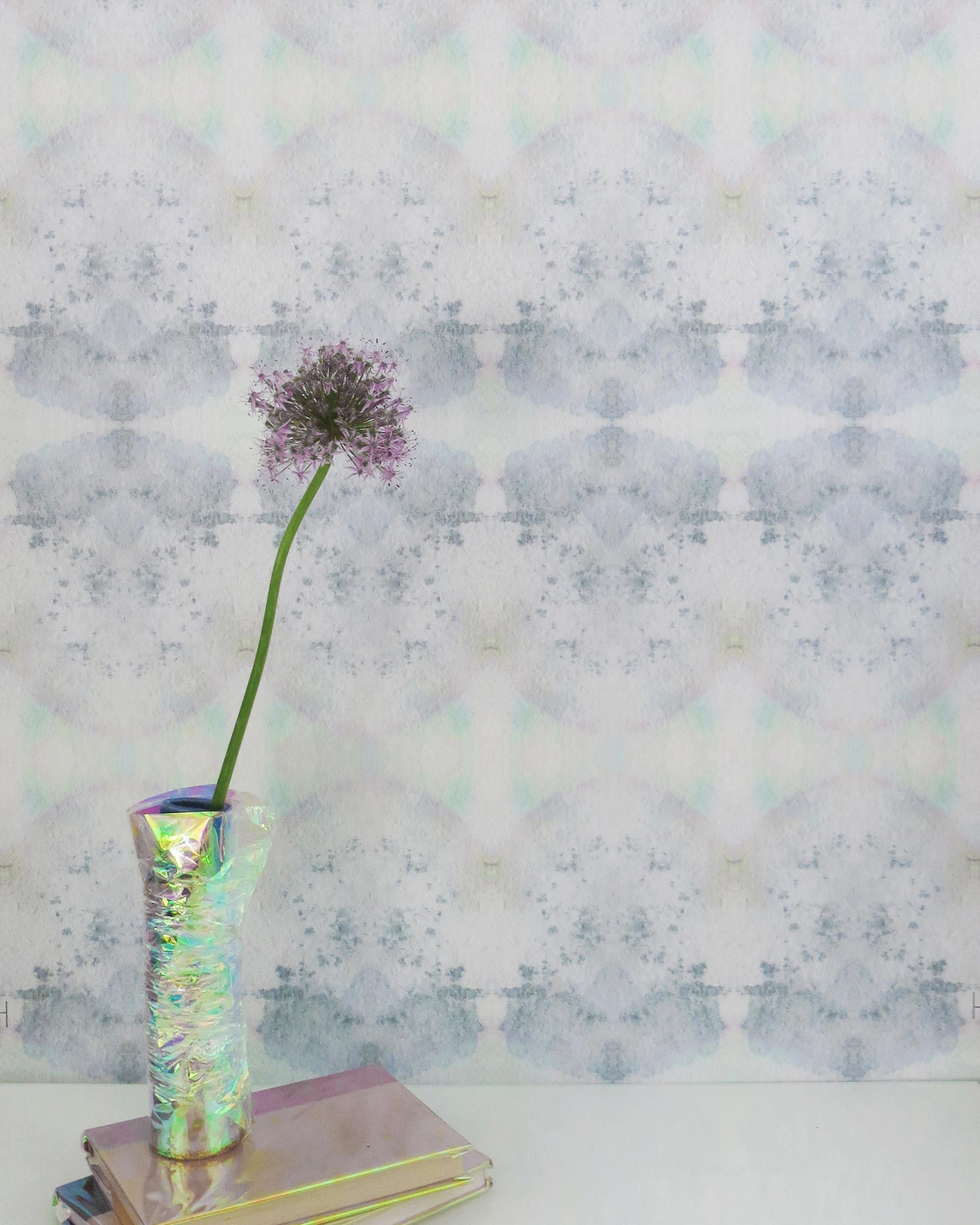 A vase with a flower stands in front of a wall papered in a watercolor ink blot pattern in shades of gray, green and pink.