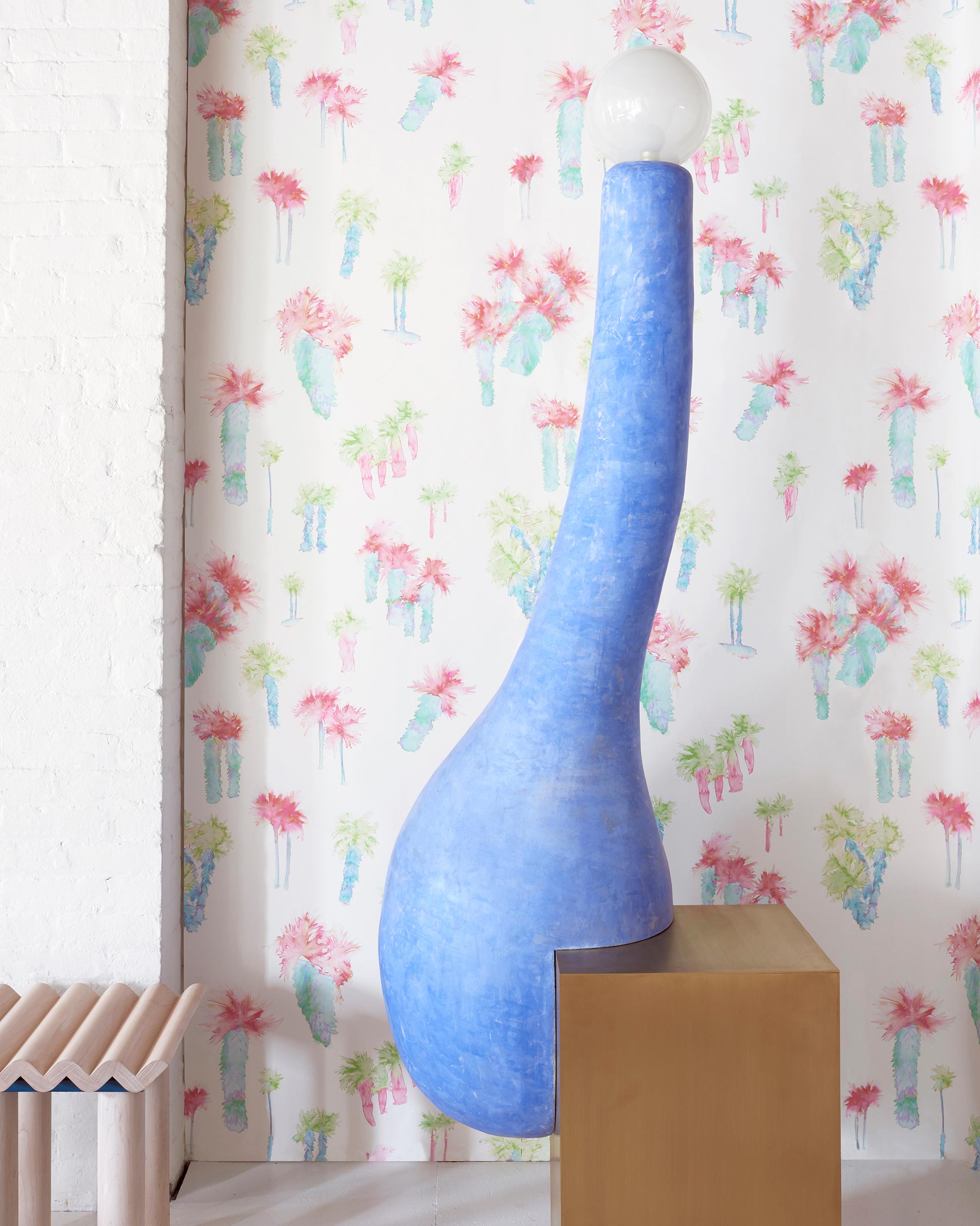 A modernist statue stands in front of a wall papered in a playful palm tree print in a rainbow of neon on a white field.
