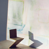 A modernist chair stands in front of a wall papered in a watercolor grid print in pastels.