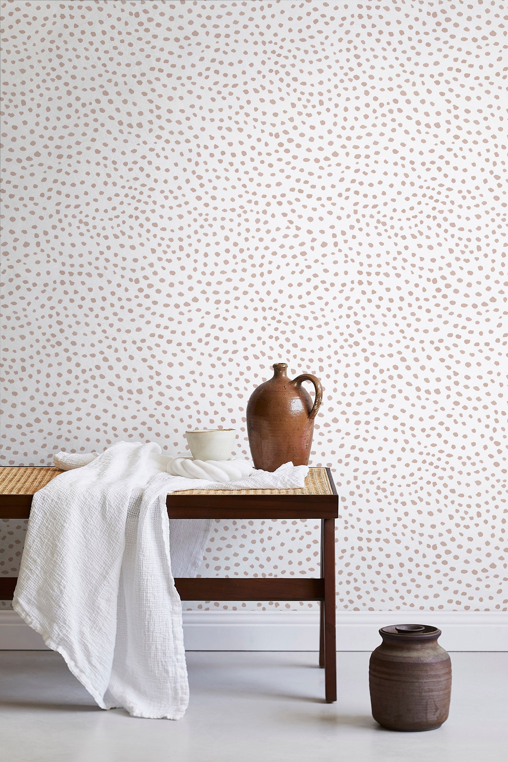 A bench with a towel and clay vases stands in front of a wall papered in a scalloped dot print in blush and white.