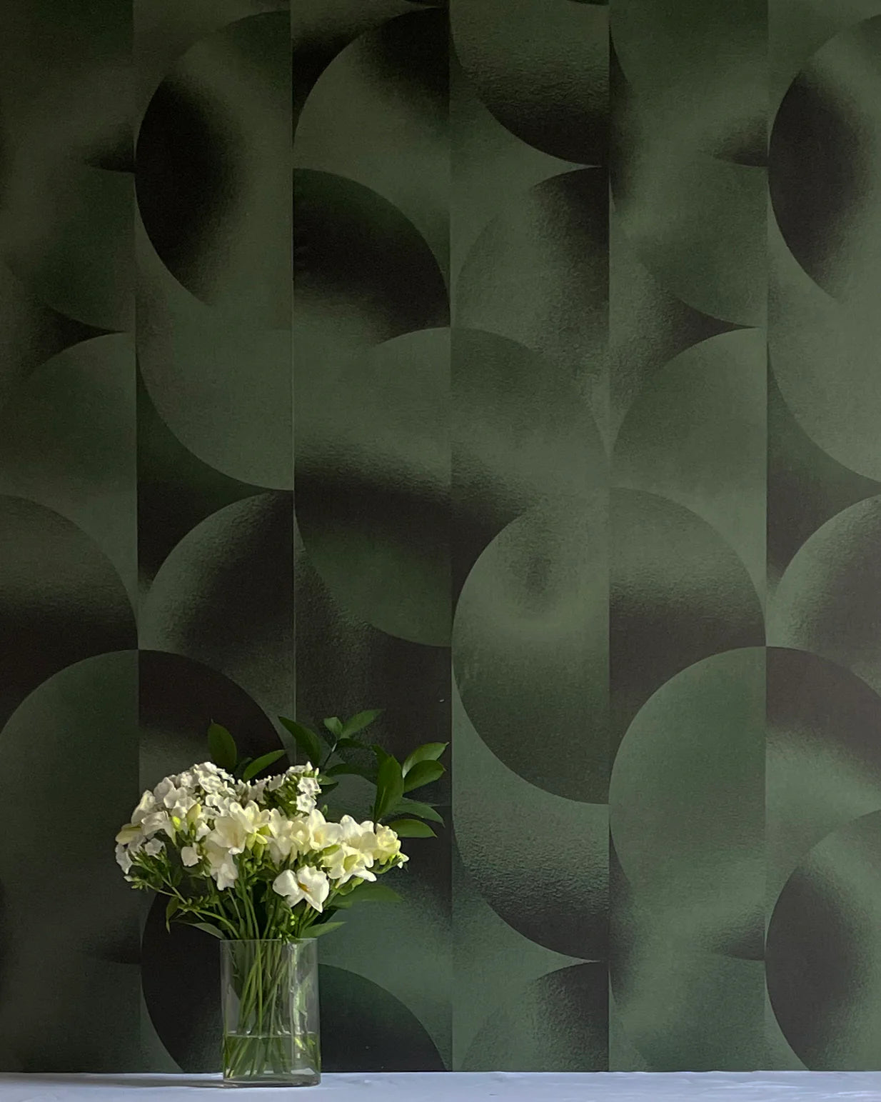 A vase of flowers stands in front of a wall covered in an abstract curvilinear print in mottled green and black.