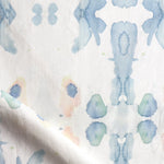 Draped fabric yardage in a watercolor ink splash print in shades of pastel blue and pink on a cream field.