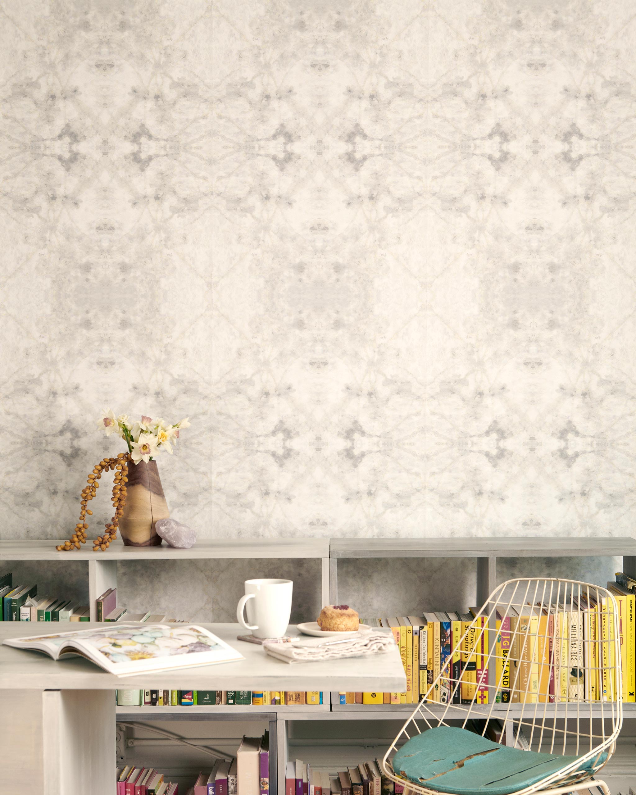 A cluttered desk and bookshelf stand in front of a wall papered in a watercolor textural print in cream, gray and tan.