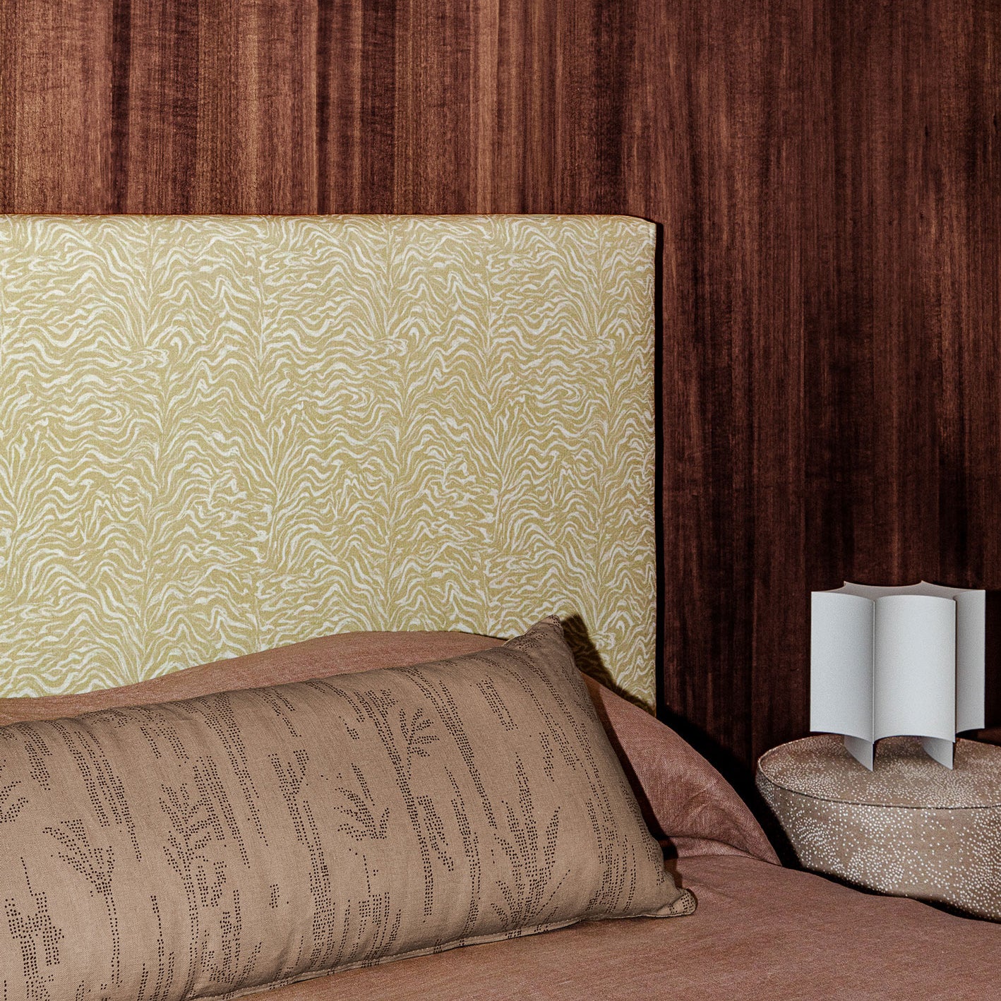 Bedroom scene with dark wood paneled wall, tan and white zebra headboard, clay colored bedding and an upholstered ottoman with white sculputral lamp