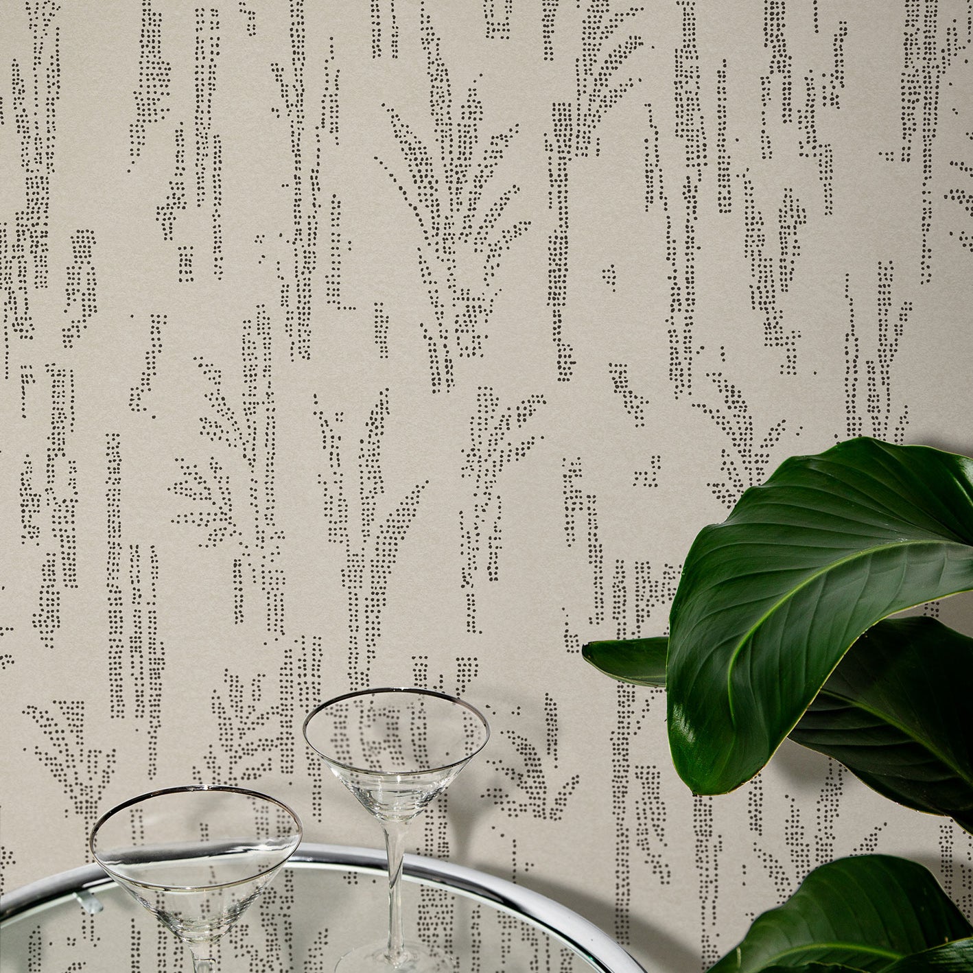 Close up of a steel and glass side table with two martini glasses and tropical plant leaves against wallpaper with a stylized wheat print in black on beige.
