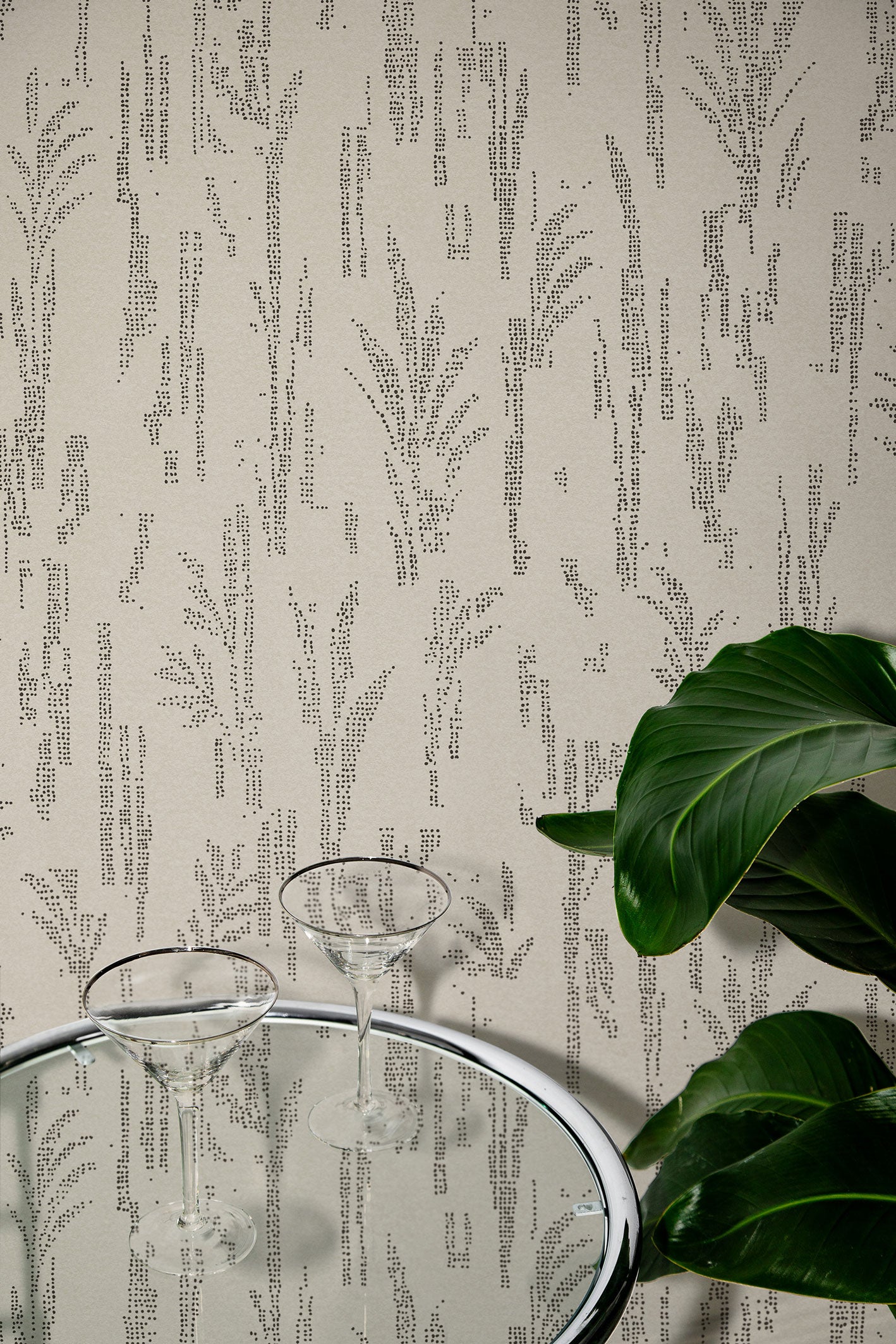 Close up of a steel and glass side table with two martini glasses and tropical plant leaves against wallpaper with a stylized wheat print in black on beige.