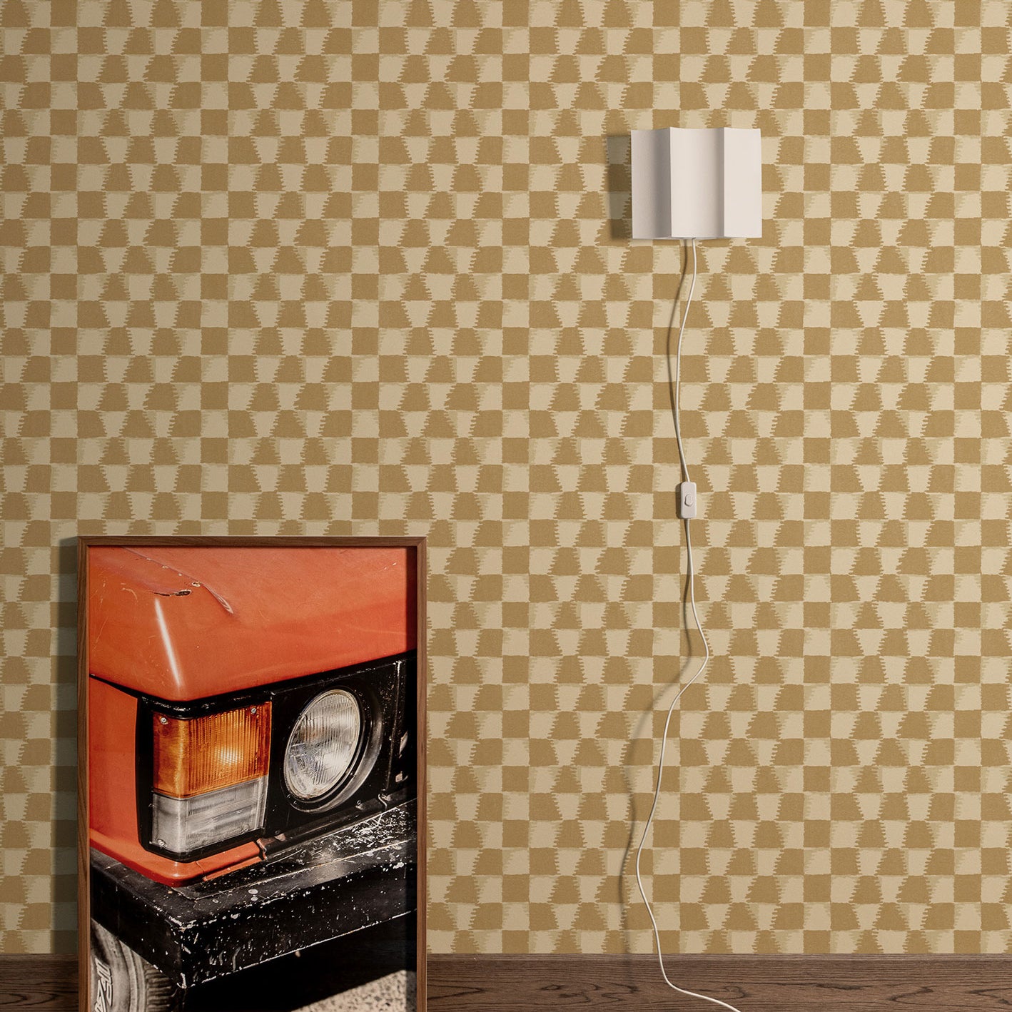 Styled interior vignette with a framed photo of a close up on an orange retro car, dark wood floors, white wall sconce with cord trailing out of the frame with a tan and beige checkerboard wallpaper backdrop.