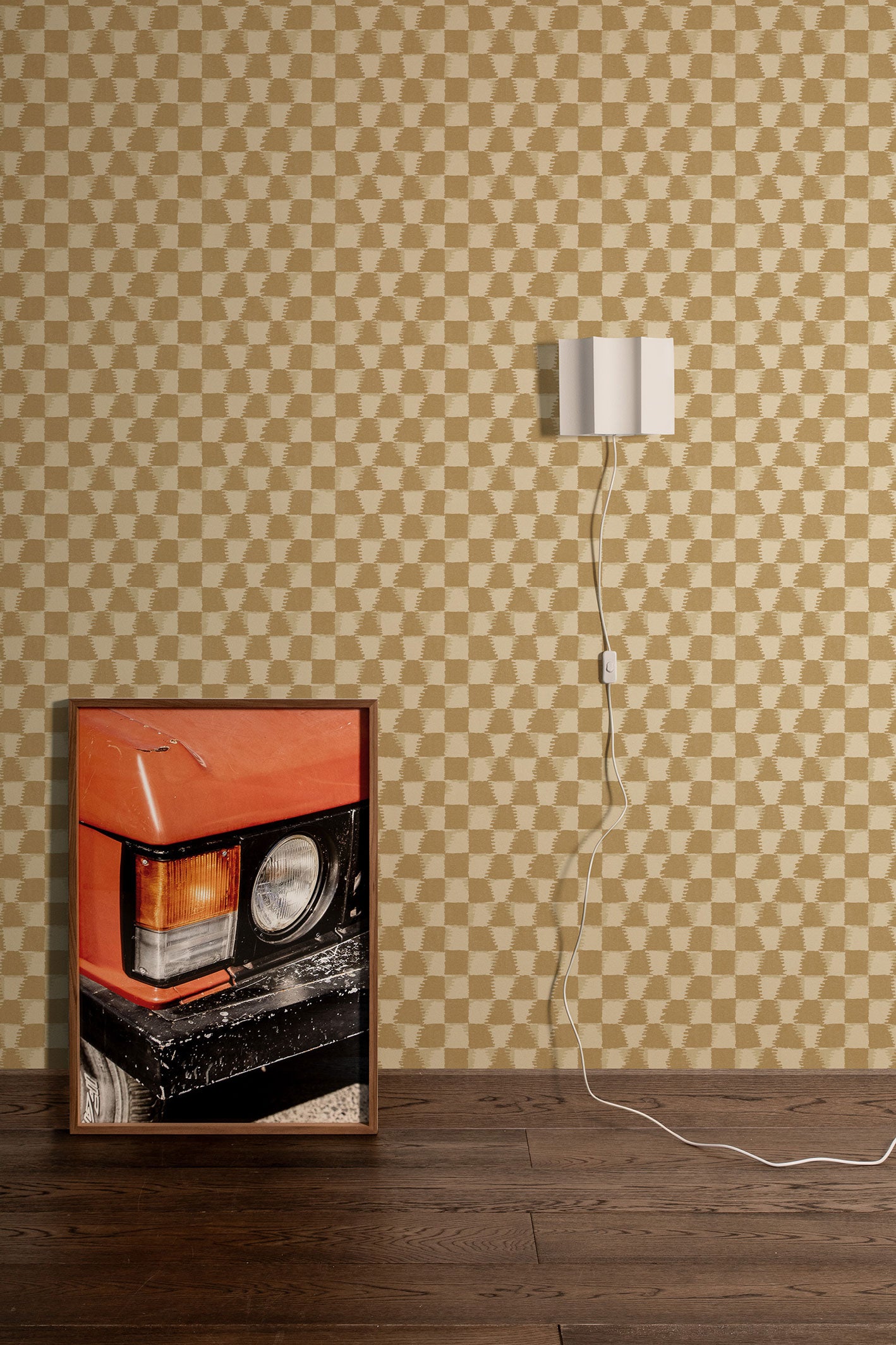 Styled interior vignette with a framed photo of a close up on an orange retro car, dark wood floors, white wall sconce with cord trailing out of the frame with a tan and beige checkerboard wallpaper backdrop.