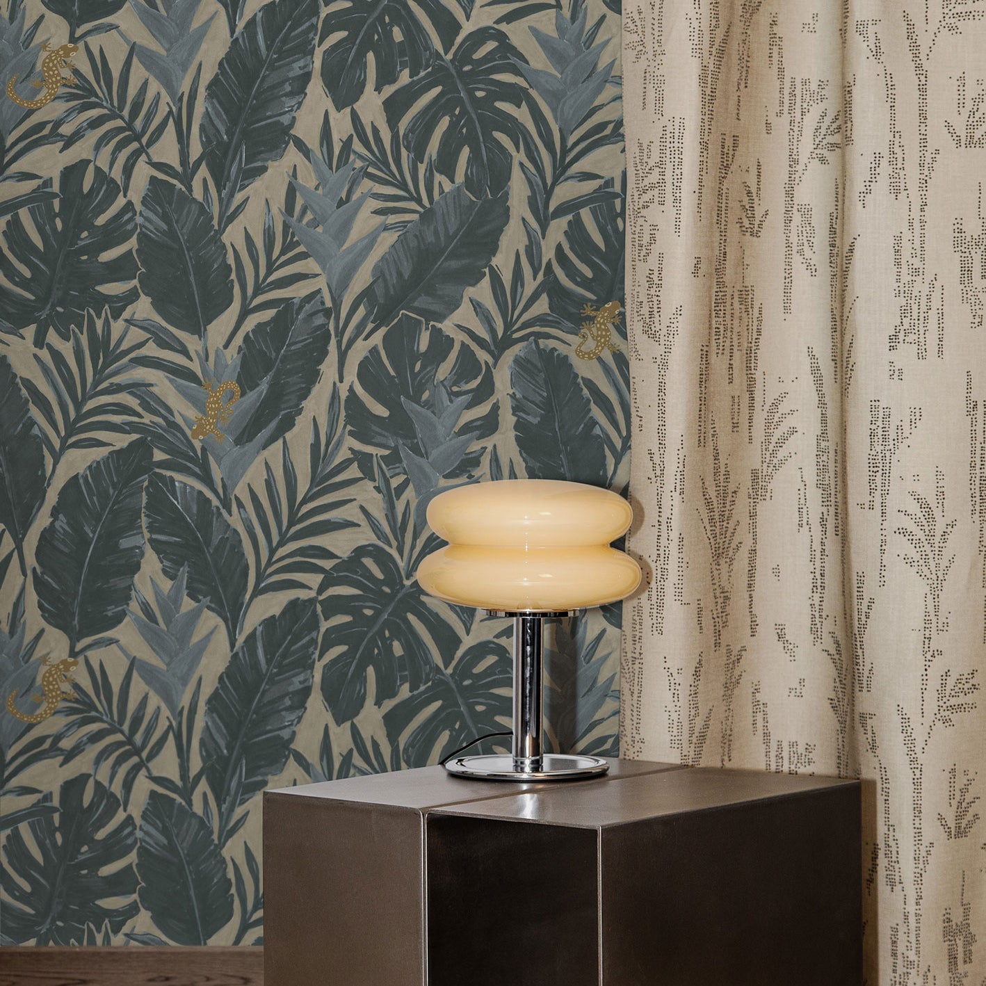 Styled interior with steel side table, retro chrome lamp with cream glass shade on a beige shag carpet against a teal and grey palm frond wallpaper and floor length curtain with a subtle dotted pattern in beige.