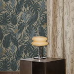 Styled interior with steel side table, retro chrome lamp with cream glass shade on a beige shag carpet against a teal and grey palm frond wallpaper and floor length curtain with a subtle dotted pattern in beige.