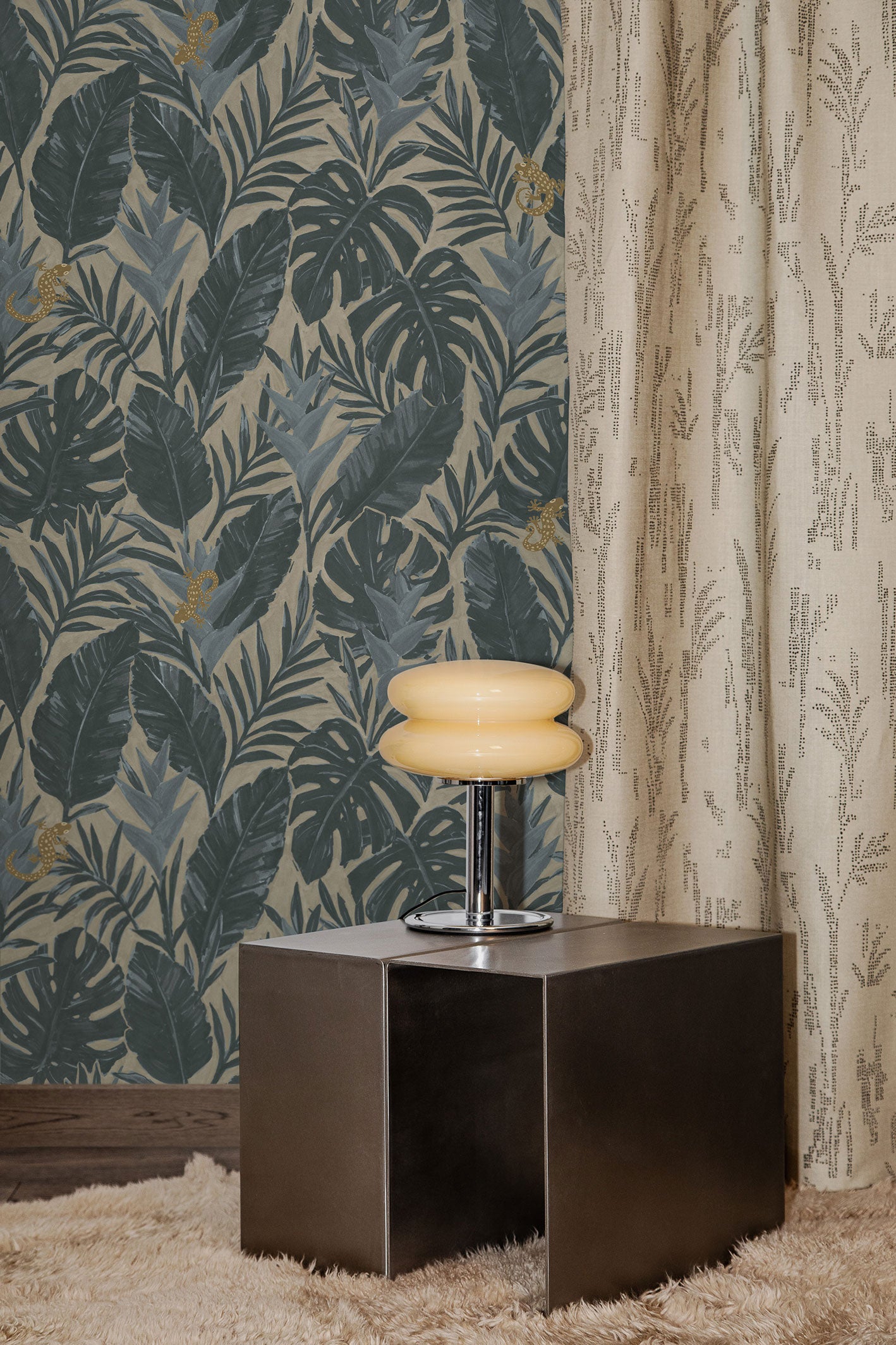 Styled interior with steel side table, retro chrome lamp with cream glass shade on a beige shag carpet against a teal and grey palm frond wallpaper and floor length curtain with a subtle dotted pattern in beige.