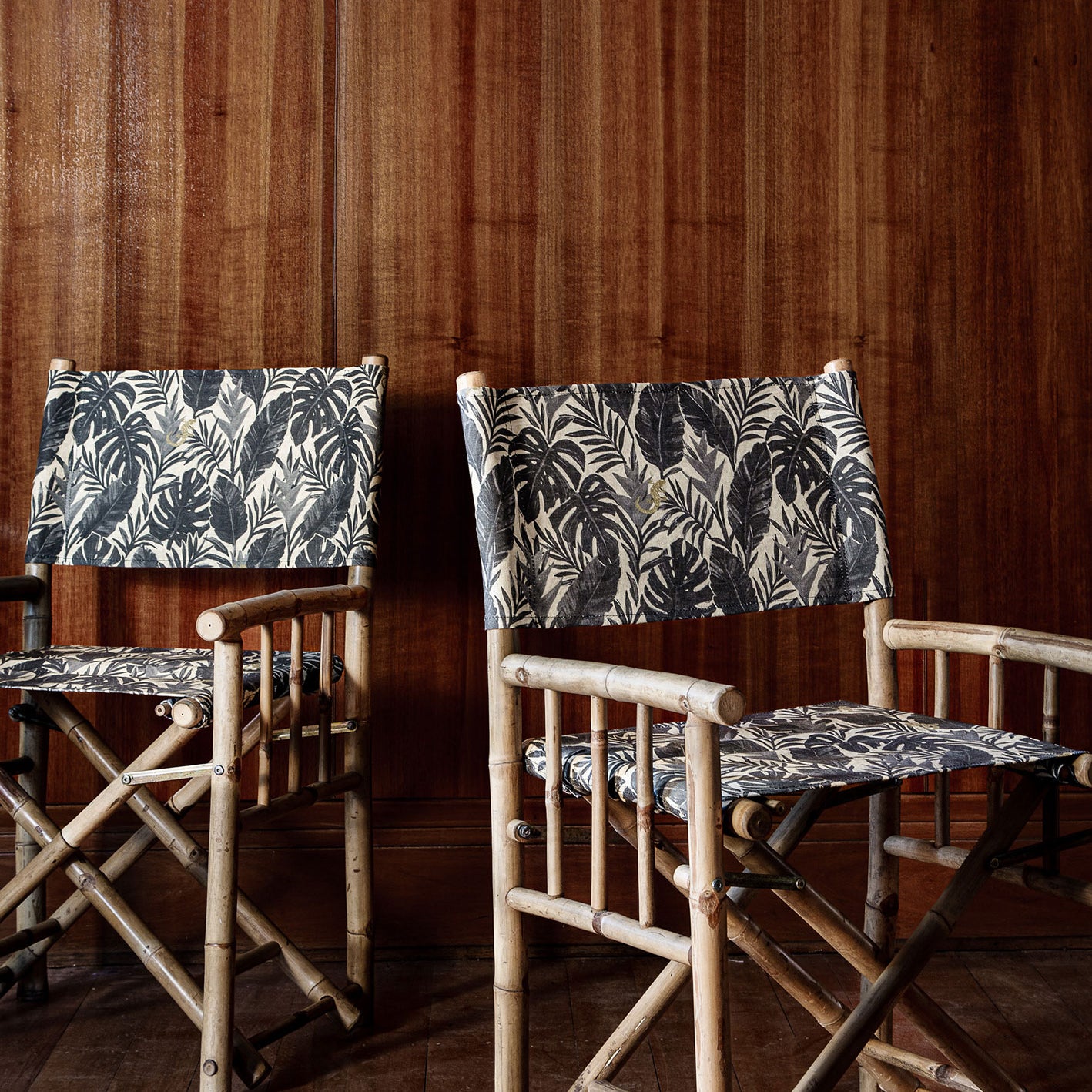 Bamboo director's chair with black and grey palm frond fabric upholstery against a dark wood paneled wall and matching floor.