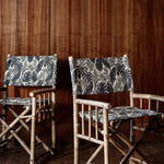 Bamboo director's chair with black and grey palm frond fabric upholstery against a dark wood paneled wall and matching floor.