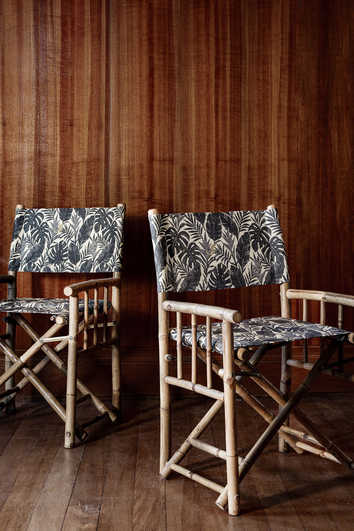 Bamboo director's chair with black and grey palm frond fabric upholstery against a dark wood paneled wall and matching floor.