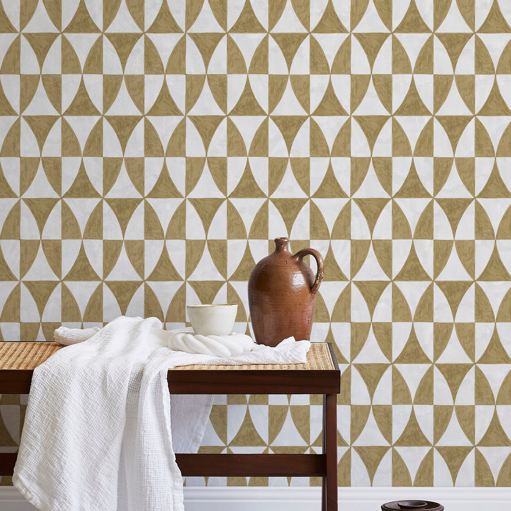 A bench with a towel and clay vases stands in front of a wall papered in a curvilinear grid print in mustard and white.