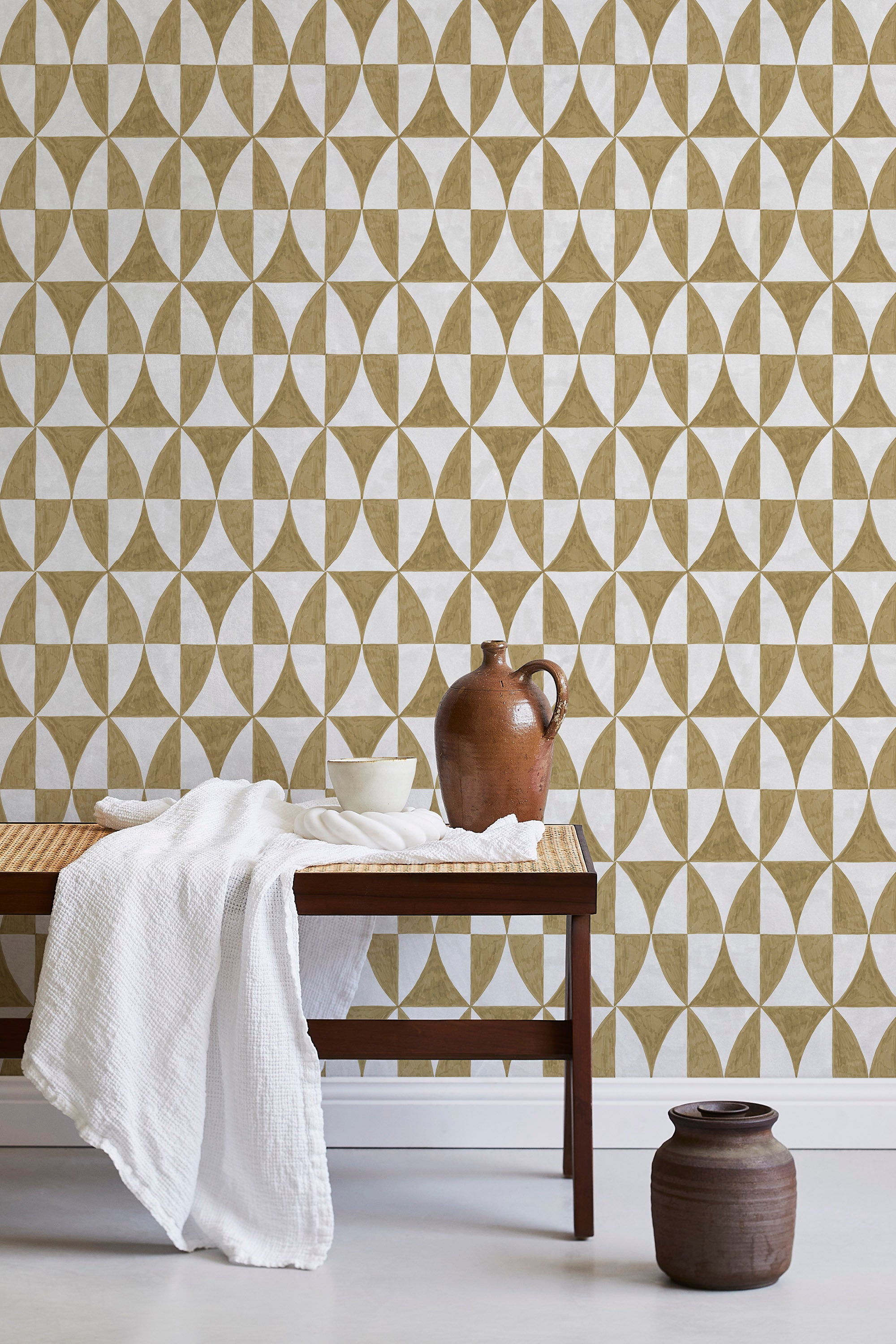 A bench with a towel and clay vases stands in front of a wall papered in a curvilinear grid print in mustard and white.