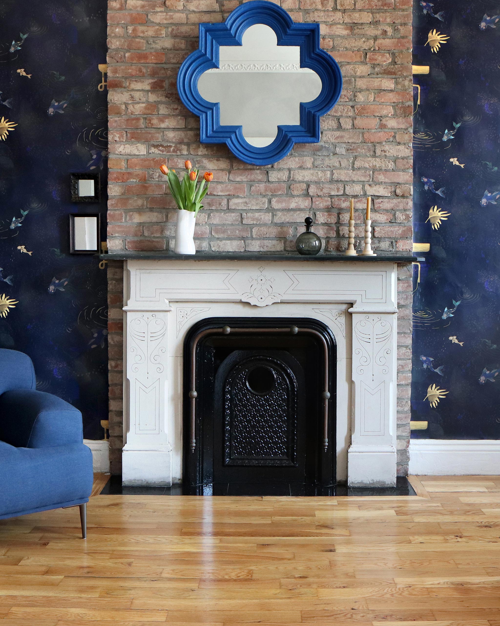 A maximalist living tableau with a fireplace and accent walls papered in a playful koi pond print in navy, gold and black.