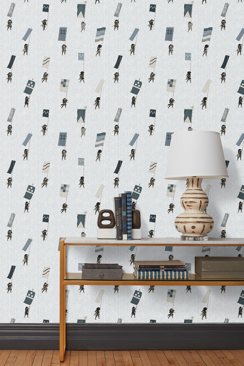 A table with books and a lamp in front of a wall papered in a pattern of Japanese soldiers carrying flags on a light gray background.