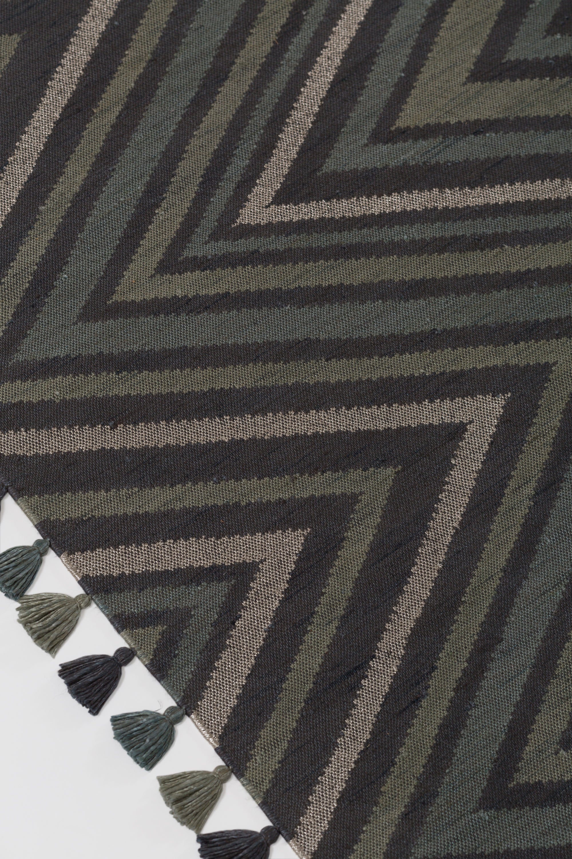 Detail of To The Point Rug in Pyrite-Metallic, a zig zag pattern in dark greens, metallic ecru and black, with a mulit color tasseled edge. 